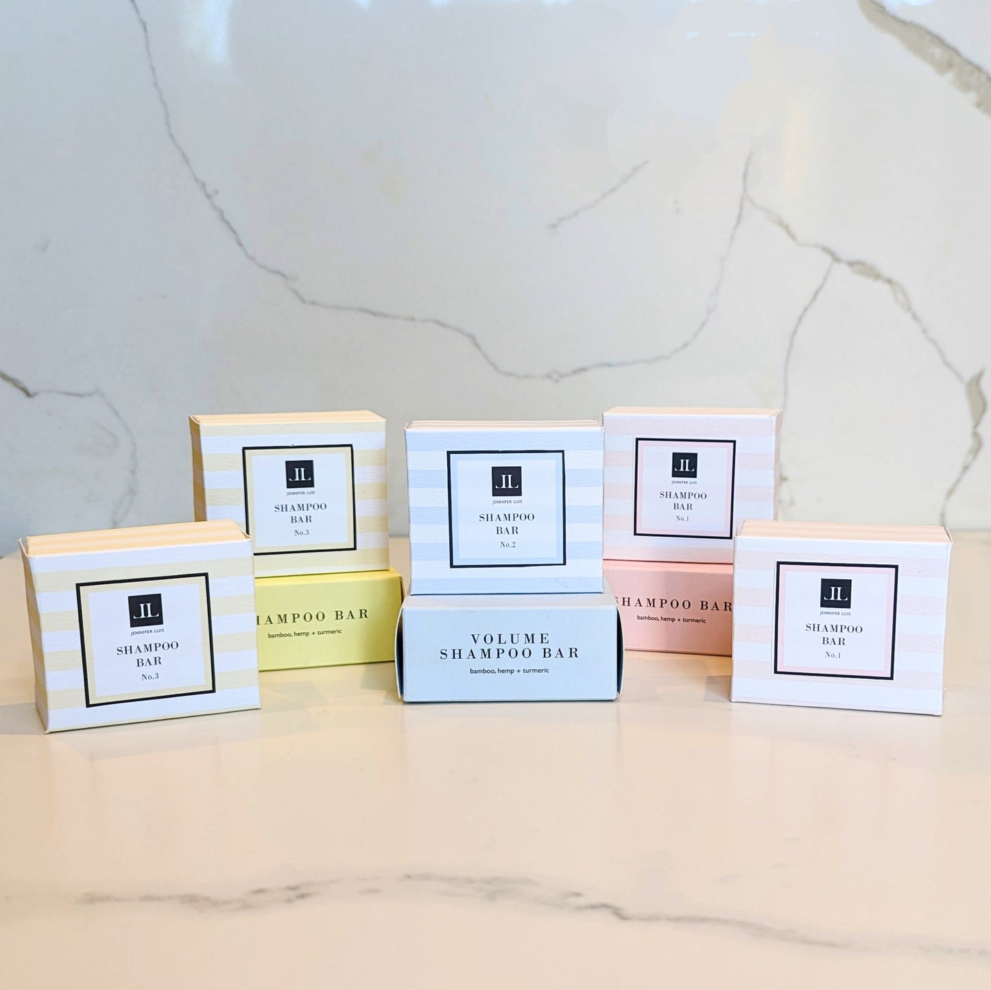 Shampoo bar boxes lined up. Two yellow striped boxes No3 50g shampoo bars, one yellow 85g shampoo bar box. Centre sits one blue box and blue and white striped box on top, the No2 85g shampoo bar box and the 50g No2 shampoo bar box. Right 3 boxes one at the bottom pink, the No1 Shampoo bar 85g box, two pink and white striped boxes the No1 50g shampoo bar boxes