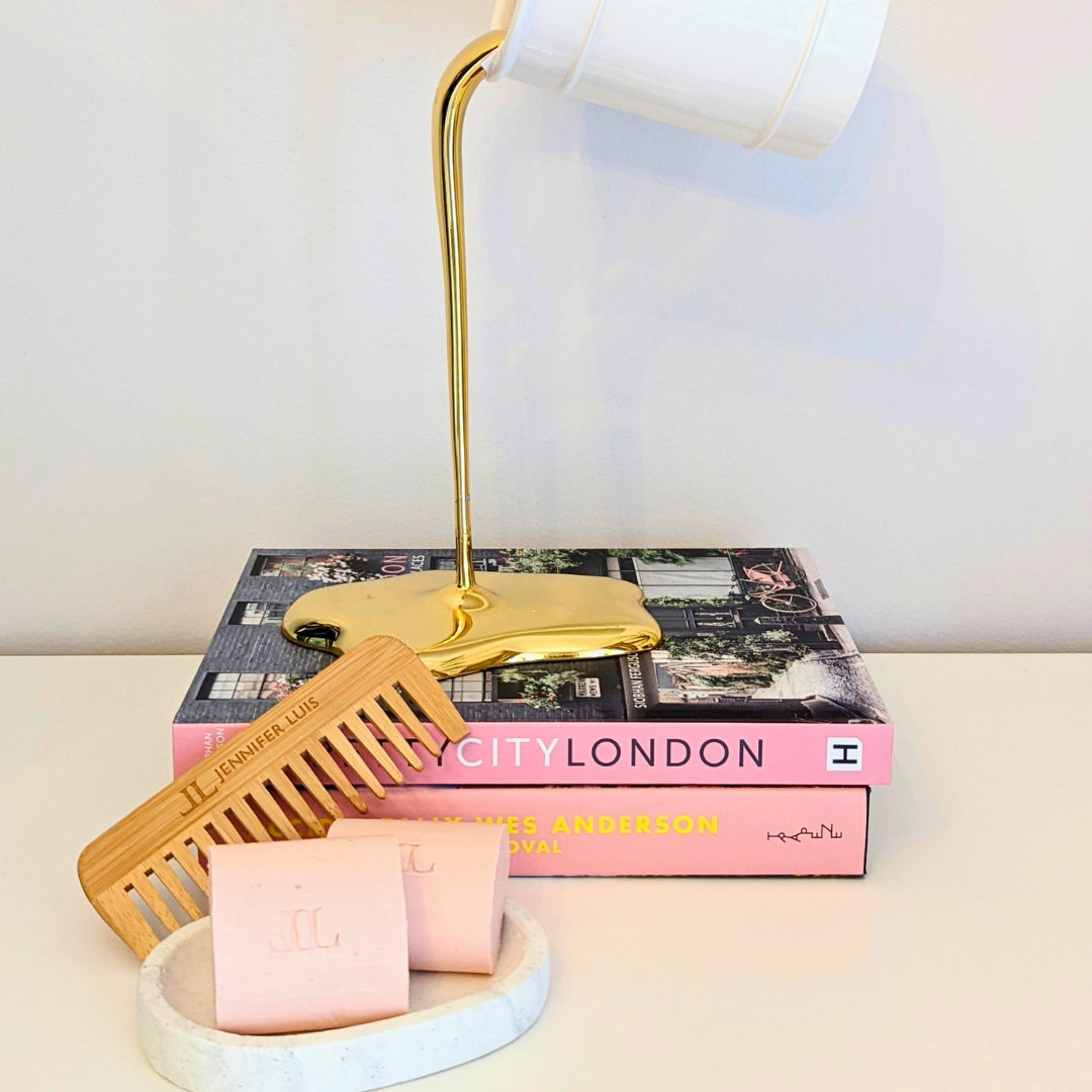 two shampoo bars for dry damaged frizzy curly hair in a soap dish with a bamboo comb propped up against them. in the background there are two pink books and a pot of paint with gold paint pouring out
